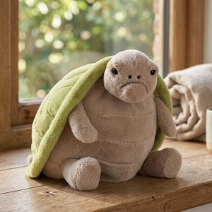 Könnte beinhalten: Ein Plüschtier in Form einer Schildkröte mit hellgrünem Panzer und beigem Körper. Die Schildkröte hat einen traurigen Gesichtsausdruck. Sie sitzt auf einer Holzoberfläche. Das Spielzeug ist weich und kuschelig.