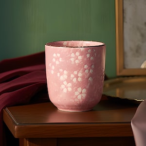 Puede incluir: Una taza de cerámica rosa con un estampado floral blanco. La taza tiene un borde oscuro y está colocada sobre una superficie de madera. El fondo incluye una pared verde, un espejo y tela drapeada.