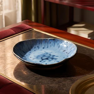 May include: A shallow, round ceramic bowl featuring a reactive glaze with a gradient of light blue and dark blue tones. The bowl has an irregular, organic rim and a smooth, glossy finish that creates a mottled, speckled appearance across the surface.