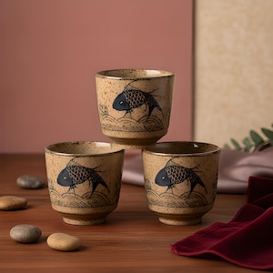 May include: Three ceramic tea cups with a speckled beige finish, each featuring a hand-painted black fish illustration swimming above stylised waves. The cups have a cylindrical shape with a slightly tapered base and a smooth, glazed texture.