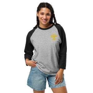 Könnte beinhalten: Ein Baseball-Shirt mit einem meliert grauen Rumpf und schwarzen Dreiviertelärmeln. Das Oberteil zeigt ein kleines, gesticktes gelbes Baummotiv auf der oberen linken Brustseite, das einen detaillierten Stamm und eine verzweigte Krone darstellt.