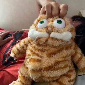 May include: A plush orange and tan cat toy with large green eyes and white whiskers. The stuffed animal has a striped pattern and is being held up by a hand.