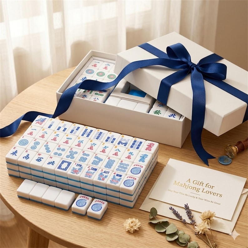 May include: A complete Mahjong set with white tiles featuring blue and red symbols, presented in a white gift box tied with a navy blue ribbon. The set includes a card that reads "A Gift for Mahjong Lovers".