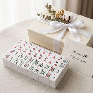 May include: A complete Mahjong set with white tiles featuring red and green symbols, arranged in front of a cream-colored gift box tied with a white ribbon. A card reads "Mahjong Gift, Enjoy the game!"