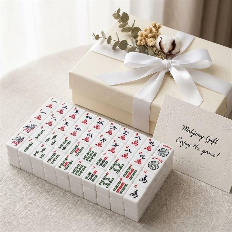 May include: A complete Mahjong set with white tiles featuring red and green symbols, arranged in front of a cream-colored gift box tied with a white ribbon. A card reads "Mahjong Gift, Enjoy the game!"