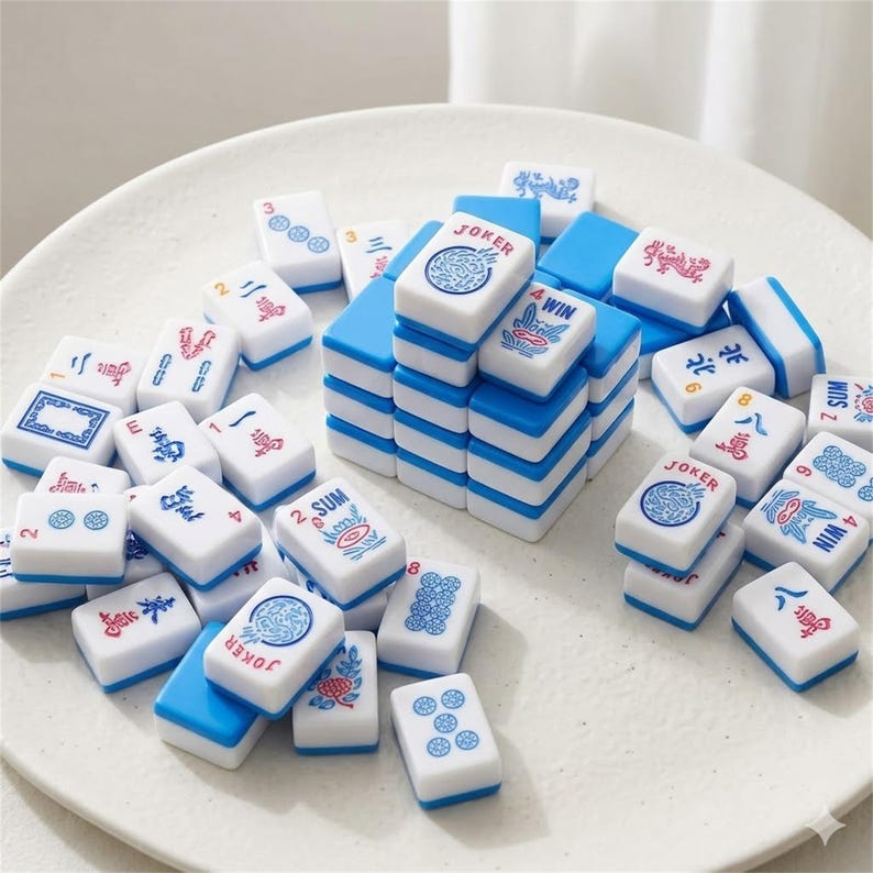 May include: A collection of white and blue Mahjong tiles. The tiles have various symbols, numbers, and Chinese characters in red and blue. Some tiles are stacked, while others are scattered on a white surface.