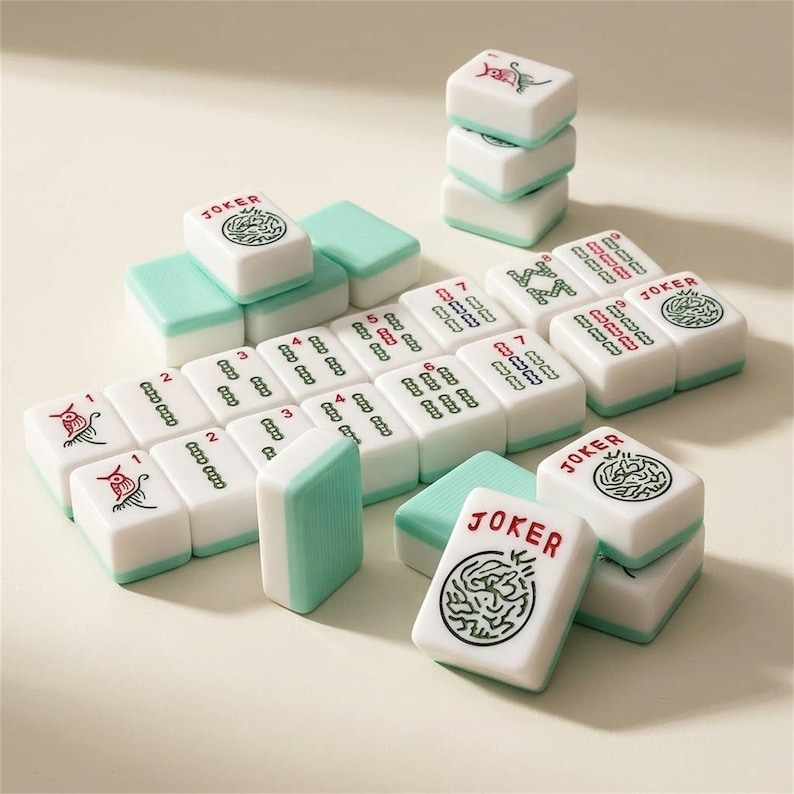 May include: A set of white and teal Mahjong tiles. The tiles have red and green symbols and numbers. Some tiles are stacked, and others are arranged in rows. The word "JOKER" is printed on some tiles.