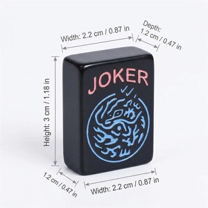 Może przedstawiać: Czarny prostokątny kafelek do gry ze słowem "JOKER" w kolorze r&oacute;żowym i niebieskim wzorem. Płytka ma 3 cm wysokości, 2,2 cm szerokości i 1,2 cm głębokości. Element gry do gier planszowych.