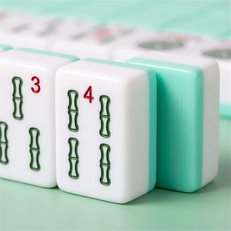 May include: Close-up of white Mahjong tiles with green bamboo symbols and red numbers. The tiles have a light green border. The number "3" is on one tile, and the number "4" is on another. The background is a light green color.