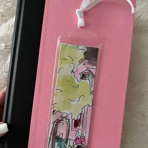 May include: A clear plastic bookmark with a watercolor illustration of a pink house with a green tree and flowers. The bookmark is attached to a white ribbon.