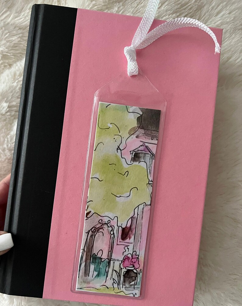 May include: A clear plastic bookmark with a watercolor illustration of a pink house with a green tree and flowers. The bookmark is attached to a white ribbon.