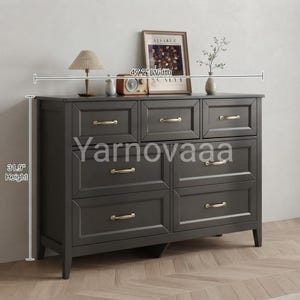 May include: A dark gray dresser with seven drawers, each featuring a metallic handle. The dresser measures 47.2 inches in width and 31.9 inches in height. The top surface holds a lamp, a radio, a framed picture, and a small vase with greenery.