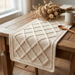 May include: Cream-colored, hand-crocheted table runner with a diamond pattern. The runner is placed on a wooden table, alongside a mug, wooden spoon, and dried flowers. The texture of the crochet is visible.