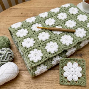 Può includere: Una coperta all'uncinetto verde salvia con motivi floreali bianchi in un motivo a quadrati della nonna. Un uncinetto è appoggiato sulla coperta. Sono visibili anche due gomitoli di lana e un sottobicchiere abbinato. La coperta misura circa 100 cm x 100 cm.