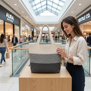 May include: A tri-color tote bag with a cream, gray, and dark gray striped design. The bag has two handles and is displayed on a wooden surface in a shopping mall setting. The bag is made of a woven material.