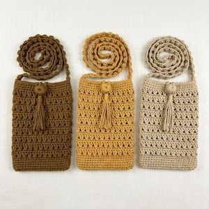 Op de afbeelding: Drie gehaakte crossbody tassen in bruin, mosterdgeel en beige. Elke tas heeft een lange, spiraalvormige band, een kwast en een decoratieve knoop. De tassen zijn rechthoekig met een textuur, open geweven ontwerp.