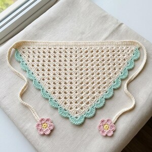 May include: Cream-colored crochet bandana with a scalloped edge in light blue. The bandana has a triangular shape with two cream-colored ties, each adorned with a pink and yellow flower. The open weave design adds a delicate touch.
