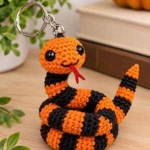 Può includere: Un portachiavi a forma di serpente all'uncinetto con strisce orizzontali alternate di filato arancione brillante e nero. Il serpente ha due grandi occhi rotondi e neri lucidi e una piccola lingua biforcuta in feltro rosso.