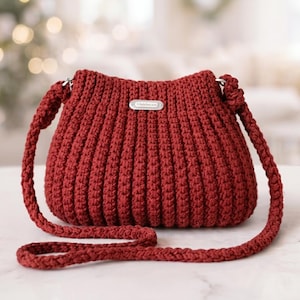 May include: A burgundy crochet handbag with a long, braided strap. The bag features a ribbed texture and a small, silver-toned label on the front. The strap is attached to the bag with silver-toned hardware. The background is blurred, suggesting a soft focus.