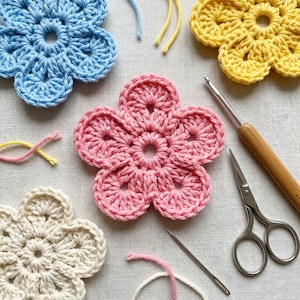 Puede incluir: Flores de ganchillo hechas a mano en varios colores, incluyendo azul, rosa, amarillo y crema. La imagen también muestra herramientas de ganchillo como un ganchillo, tijeras y una aguja, junto con restos de hilo. Las flores están dispuestas sobre una superficie neutra.
