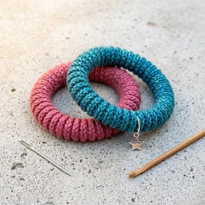 May include: Two handmade, knitted bracelets in teal and pink, resting on a concrete surface. The teal bracelet has a silver star charm. A needle and a crochet hook are also present, suggesting a craft project.