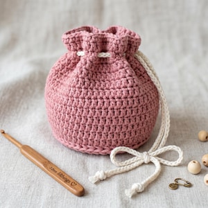 Può includere: Un sacchetto all'uncinetto rosa polveroso con coulisse e fiocco color crema. Un uncinetto di legno con la scritta "3.0mm lilthings" è accanto alla borsa. La borsa è rotonda e ha una parte superiore arricciata.