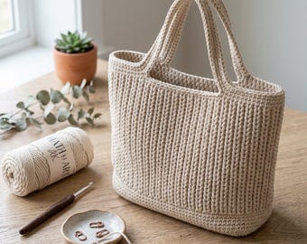 May include: A cream-coloured, hand-crocheted tote bag with two handles. The bag has a ribbed texture and is made of thick yarn. A ball of yarn, a crochet hook, and a small dish with stitch markers are also visible.