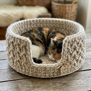 May include: A beige, hand-crocheted cat bed with a sleeping calico cat inside. The bed has a textured, woven design and a small wooden tag that reads "CAT'S CACHE". The bed is on a wooden surface.