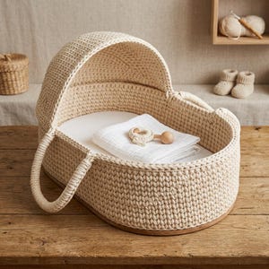 May include: A beige crocheted baby bassinet with a curved hood and handles. Inside, a white blanket and a wooden and crocheted rattle rest on a white mattress. The bassinet sits on a wooden base, suggesting a handmade nursery item.