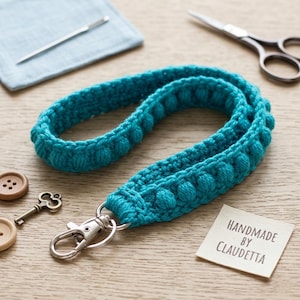 May include: A handmade teal crochet lanyard with a silver-colored clasp. The lanyard features a textured design with bobbles. The image also shows a pair of scissors, a needle, and a tag that reads "Handmade by Claudetta."