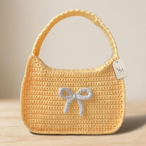 May include: A handmade, yellow crochet handbag with a matching handle. A light blue bow is attached to the front. A small tag with the letters "VM" hangs from the handle. The bag is a soft, textured material.