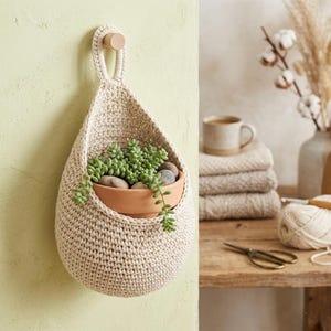May include: A cream-colored, crocheted hanging planter with a wooden dowel for hanging. A terracotta pot filled with green succulents and small stones sits inside the planter. The planter is hanging on a light green wall.