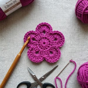 Può includere: Primo piano di un fiore magenta all'uncinetto con un uncinetto di legno, forbici e un ago da lana. Sono visibili anche due gomitoli di filato magenta e un'etichetta con la scritta "5.0mm (H) WORSTED". Gli oggetti sono su una superficie grigio chiaro.