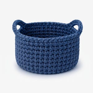 Puede incluir: Una cesta de almacenamiento redonda y profunda hecha de un cordón grueso de color azul oscuro con un patrón de punto de ganchillo denso. La cesta cuenta con dos asas integradas y resistentes en los lados opuestos, creando una textura uniforme.
