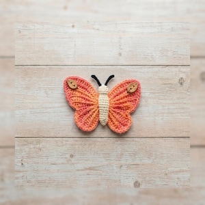 May include: A handmade crochet butterfly in shades of orange and pink, with a cream-colored body and black antennae. The butterfly's wings are adorned with small wooden buttons. The craft item is set against a light wood grain background.