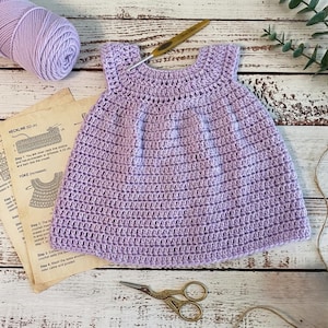 May include: A handmade, light purple crochet baby dress. The dress is sleeveless with a round neckline and a flared skirt. A ball of matching yarn, a crochet hook, scissors, and crochet instructions are also visible.