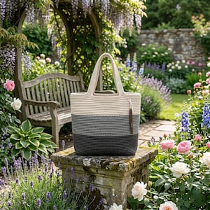 May include: A crocheted tote bag with a cream, gray, and dark gray striped design. The bag has a cream-colored handle and a brown leather tag. The bag is sitting on a stone pedestal in a garden setting.
