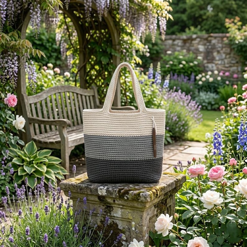 May include: A crocheted tote bag with a cream, gray, and dark gray striped design. The bag has a cream-colored handle and a brown leather tag. The bag is sitting on a stone pedestal in a garden setting.