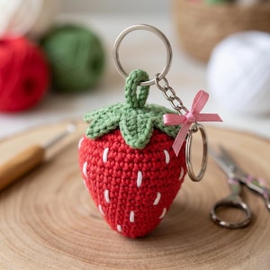 May include: A handmade crochet strawberry keychain. The red strawberry has white seed details and a green leafy top. A small pink ribbon is tied to the silver chain and keyring. The background includes yarn and a wooden surface.