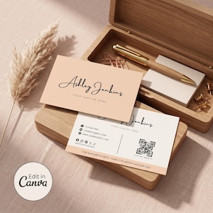 May include: Two rectangular business cards with a peach and white color scheme. The top card displays the name Ashley Jenkins in black script with the text YOUR TAGLINE HERE. The bottom card includes contact icons, a QR code, and the address 1238 Maplewood Drive, Charlotte, North Carolina (NC).