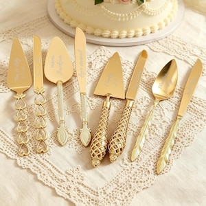 Puede incluir: Una colección de utensilios dorados para servir, que incluyen palas para pasteles, cuchillos y una cuchara, dispuestos sobre un mantel de encaje. Algunos utensilios tienen diseños decorativos de corazón, enrejado y plumas. El texto está grabado en algunas piezas.