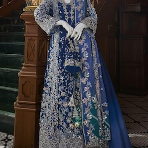May include: A formal blue gown with intricate silver embroidery and embellishments. The dress features long sleeves, a detailed bodice, and a flowing skirt. A matching blue purse and shawl complete the ensemble. The gown is displayed on a mannequin.