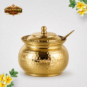 May include: A gold-colored metal container with a rounded, hammered texture body and a matching lid with a central knob. A small metal spoon is tucked into the side of the lid. The container features a polished finish and a wide base.