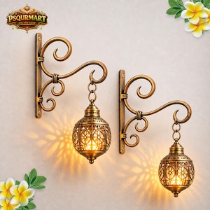 May include: Two wall-mounted lamps with an antique gold finish. Each lamp features a decorative scrollwork bracket and a spherical metal lantern with intricate floral cutouts that emit a warm, glowing light pattern onto the wall surface.