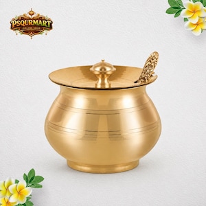 May include: A polished gold-toned brass container with a rounded body, a matching lid featuring a central knob, and a decorative spoon tucked into the side. The surface is smooth with subtle horizontal etched lines around the center.