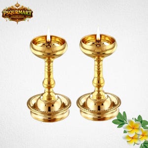 May include: Two golden oil lamps with lit wicks. The lamps have a round base, a detailed stem, and a bowl-shaped top. The lamps are made of shiny gold metal. The background is white with a logo in the top left corner and flowers in the bottom right corner.