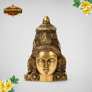 May include: A polished brass sculpture of a deity head featuring intricate engravings on the crown and large circular earrings. The face has almond-shaped eyes, a defined nose, and a calm expression, with a pointed top section of the headpiece.