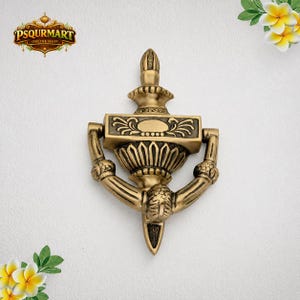 May include: A brass door knocker featuring an ornate, symmetrical design with a central urn-shaped element, a rectangular top plate with engraved floral patterns, and a curved, textured swinging handle that tapers to a pointed base.