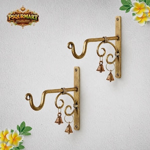 May include: Two wall-mounted metal plant brackets with a distressed gold finish. Each bracket features a curved hook for hanging items and decorative scrollwork accents. Two small, dangling brass-coloured bells are attached to each bracket.