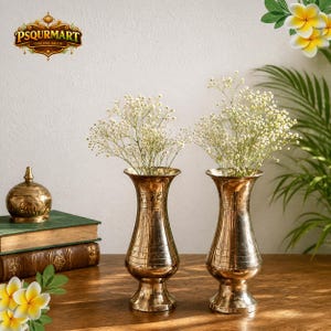 May include: Two brass vases with a fluted design, each holding a bouquet of small white flowers. The vases are displayed on a wooden surface, with a decorative brass container and books in the background. The logo "PSQURMART ONLINE SHOP" is visible.